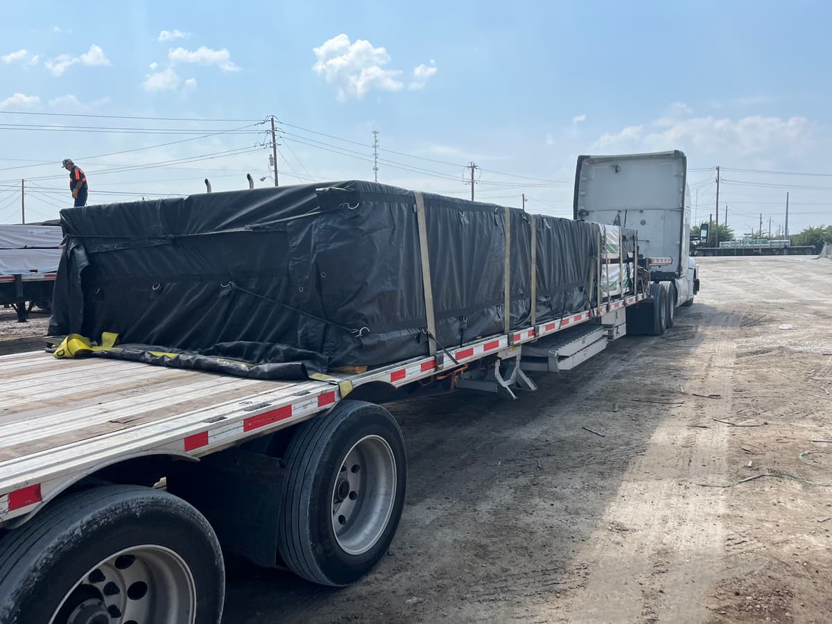 White tractor and flatbed trailer loaded with tarped, strapped freight in a gravel yard.